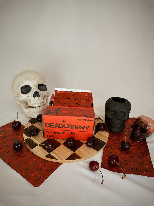 Box of 'Deadly Sweet' chocolate sheets with decorative skulls and cherries on a checkered board.